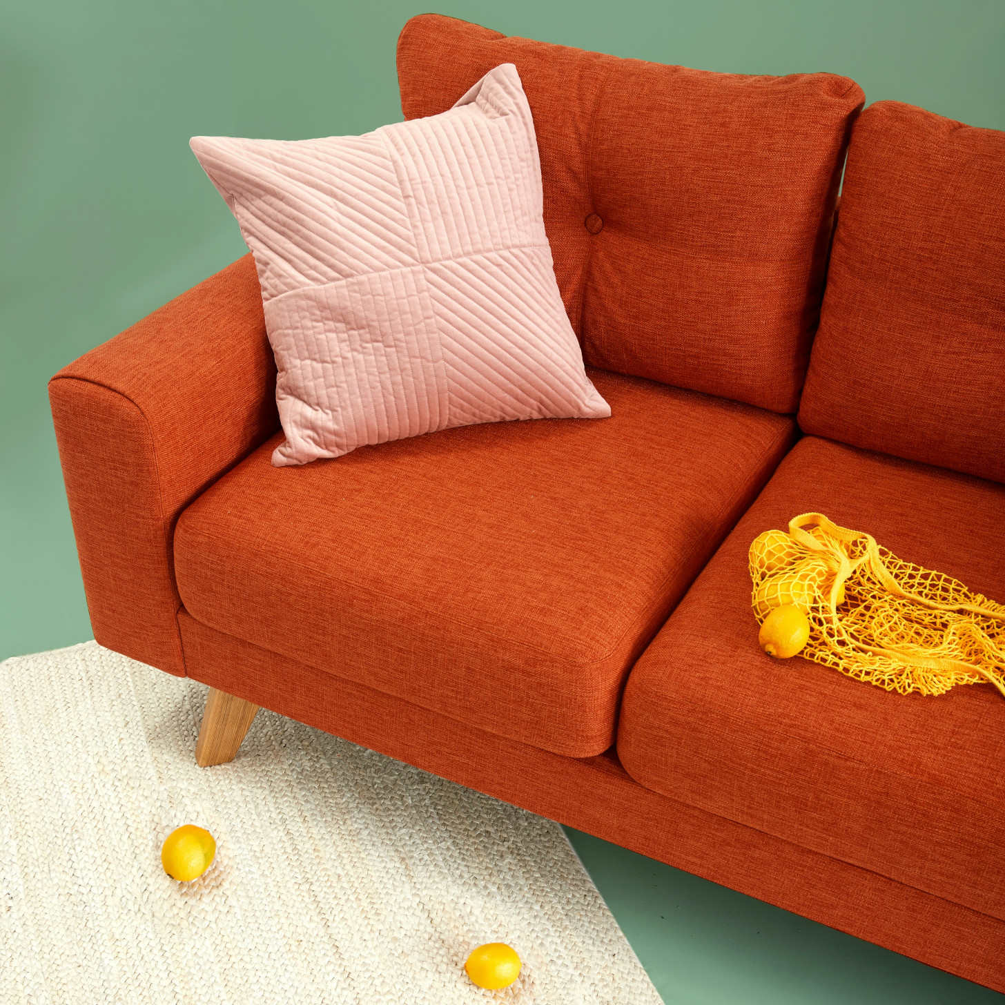 Exemplo de imagem base de um sofá vermelho com uma bolsa de limões à direita e um travesseiro decorativo no canto esquerdo. O sofá está posicionado em um ângulo, com um único pé colocado em um tapete branco. Em frente ao sofá, sobre o tapete, há dois limões.